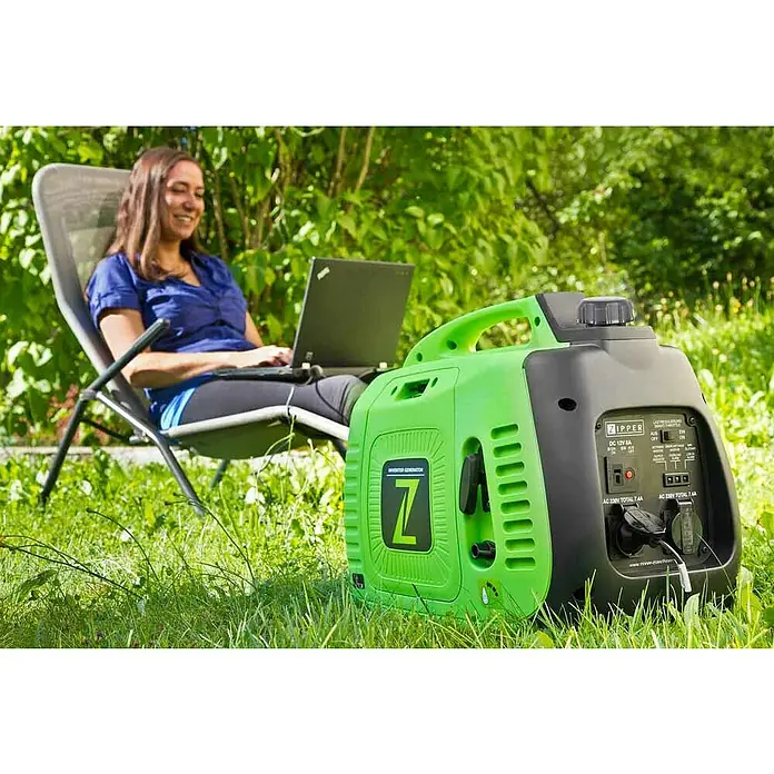 Zipper Stromerzeuger ZI-STE2000IV - 2,2 kW, Tankvolumen: 3,5 l - Ein grüner und schwarzer Zipper Inverter Generator steht im Gras, während eine Frau im Hintergrund an einem Laptop arbeitet.