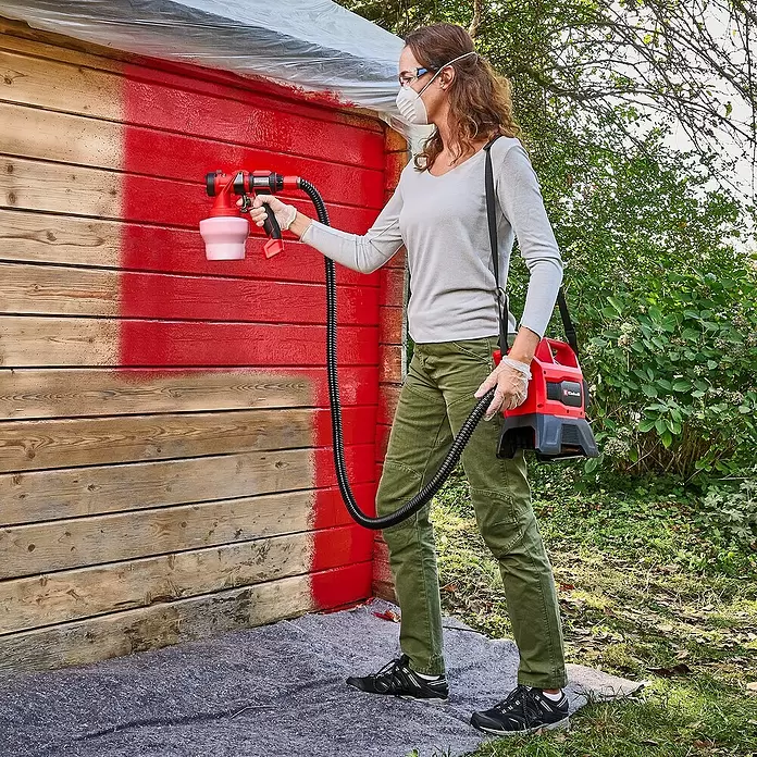 Einhell PXC 18V Sistema de pulverización de pintura de batería TE-SY 18/90 Li-Solo - 18 V, Sin batería - Mujer utiliza sistema de pulverización de pintura rojo y negro para pintar un cobertizo de madera de rojo.