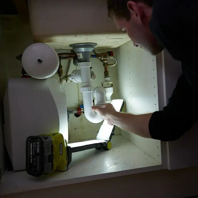 Ryobi ONE+ LED-Arbeitsleuchte - 18 V, Li-Ionen, Ohne Akku - Ein Mann untersucht ein Abflussrohr unter einem Waschbecken mit einer hellen Ryobi Arbeitsleuchte, die für gute Sicht sorgt.