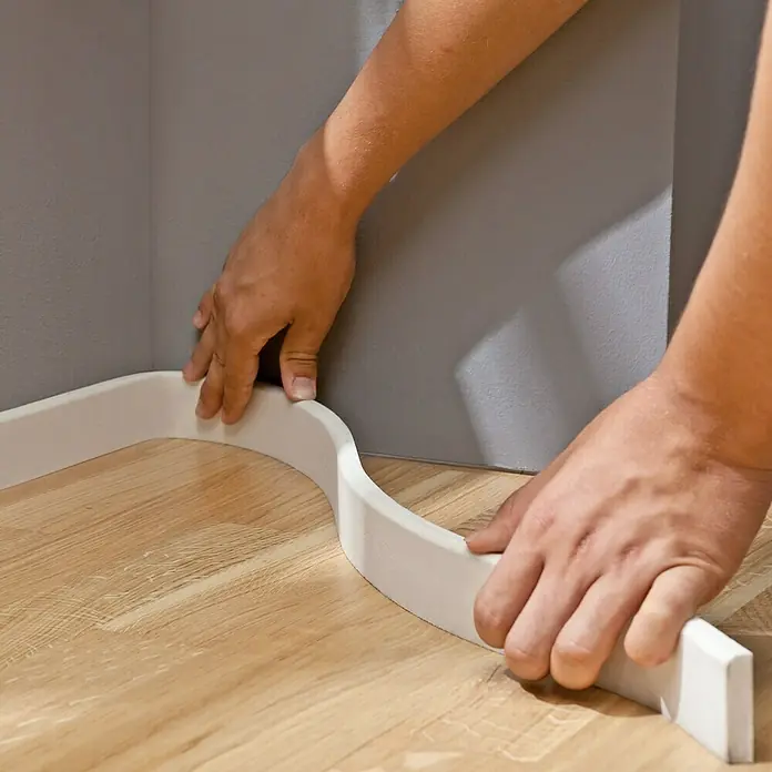 Profiles and more Sockelleiste Pro Flex - 240 cm x 10 mm x 44 mm, Weiß - Eine Person befestigt eine weiße Sockelleiste an einer grauen Wand auf einem Holzboden, um den Raum zu verschönern.