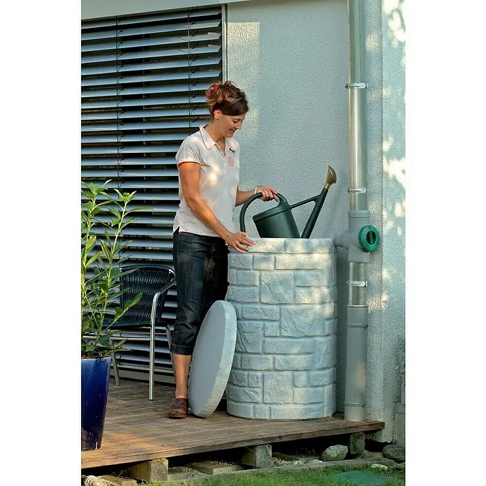Arves Regenspeicher Arcado - 230 l, Granitfarben - Frau gießt mit einer Gießkanne Wasser in einen dekorativen Regenwassertank in Steinoptik. Praktische Gartenbewässerung!