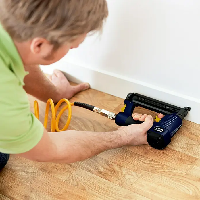 Rapid Kombidrucklufttacker AirTac PBS 121 - Klammertiefe: 12 mm - 30 mm, Geeignet für: Klammertyp 606, Pneumatisch - Nahaufnahme einer Person, die mit einem Rapid Akku-Nagler eine Fußleiste anbringt. Das Werkzeug ist ein unverzichtbarer Helfer für Heimwerker.