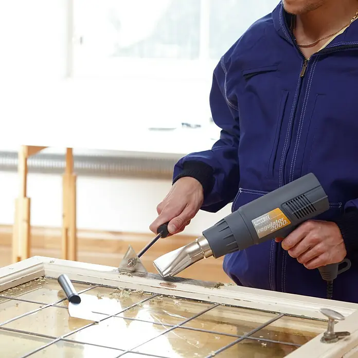 Rapid Glasschutzdüse - Breite: 50 mm, Passend für: Heißluftgebläse mit Düse Ø 35,5 mm - Ein Handwerker verwendet eine Rapid Regulator 2000 Heißluftpistole, um Farbe von einem alten Fensterrahmen zu entfernen. Das Werkzeug erhitzt die Oberfläche für die Renovierung.