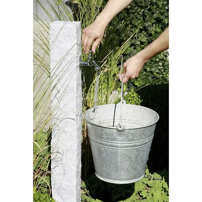 Arves Wasserzapfstelle Natura - Granitfarben, Höhe: 98 cm - Ein Mann befüllt einen Zinkeimer mit Wasser aus einem Gartenwasserhahn, der an einem Steinträger befestigt ist.