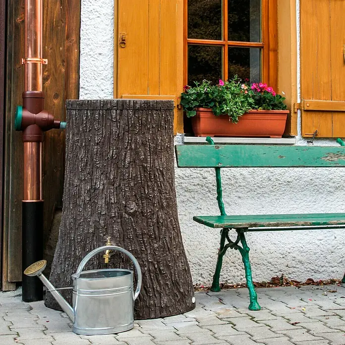 Regenspeicher Little Tree - 250 l, Dunkelbraun - Regenwassertank in Baumstammoptik mit Gießkanne und einer grünen Bank vor einem Haus.