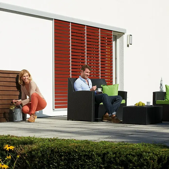 Garantia Exklusive Line Regenspeicher Woody - 350 l, Darkwood - Eine Frau gießt Pflanzen im Garten, während ein Mann auf einem schwarzen Rattansofa sitzt und Kaffee trinkt.