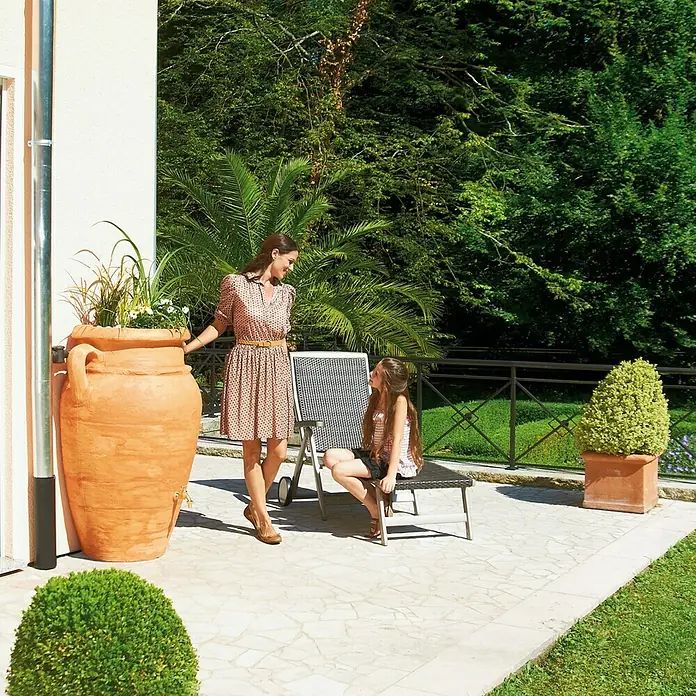 Garantia Exklusive Line Regenspeicher Amphore - 250 l, Terrakotta - Dekorative Terrakotta-Amphore als Regenwassertonne mit Bepflanzung, neben einer Frau und einem Kind auf der Terrasse.