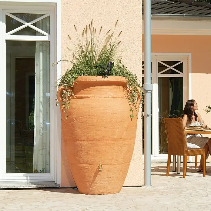 Garantia Exklusive Line Regenspeicher Amphore - 360 l, Terrakotta - Terrakottafarbener Wasserspeicher in Amphorenform mit Bepflanzung, der neben einer Hauswand auf einer Terrasse steht.