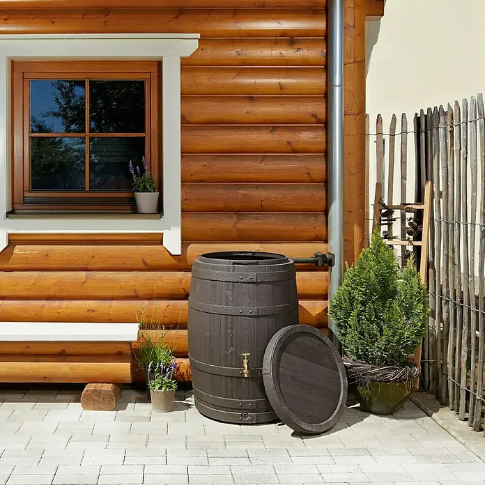Garantia Classic Line Regenspeicher Barrica - 260 l, Dunkelbraun - Ein rustikaler Regenspeicher in Fassform mit offenem Deckel steht vor einem Holzhaus, ideal für Gartenbewässerung und umweltbewusste Wassernutzung.