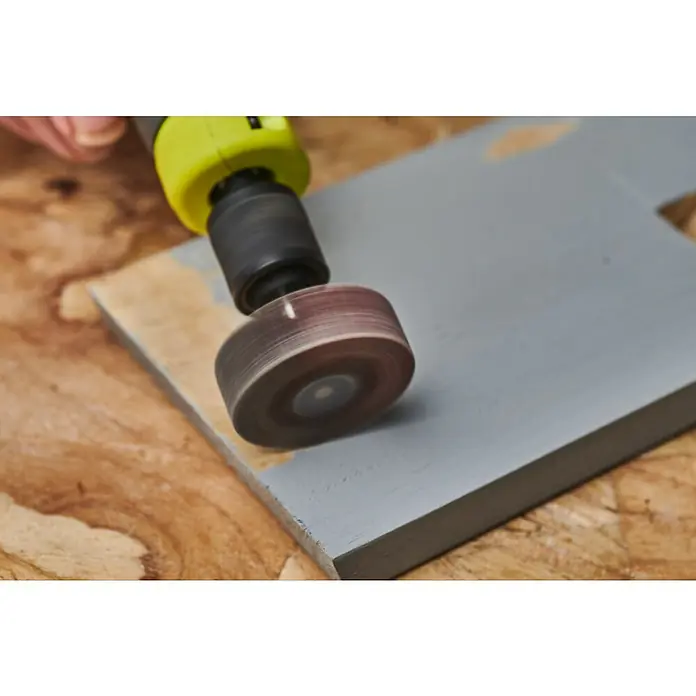 Ryobi Fächerschleifer RAR605 - Arbeitsdurchmesser: 30 mm, Körnung: 120 - Das Bild zeigt einen Schleifaufsatz an einem Bohrer, der zum Abschleifen einer grauen Holzoberfläche verwendet wird, um sie für weitere Bearbeitungsschritte vorzubereiten.