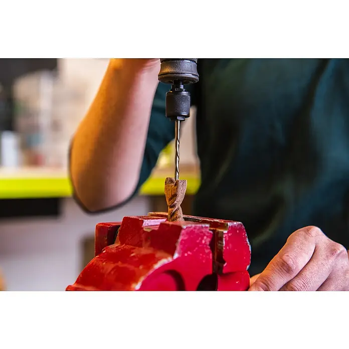 Ryobi Bohrersatz RAR404-4 - 4 Stk. - Ein Handwerker bohrt mit einer Bohrmaschine ein Loch in ein Holzstück, das in einem roten Schraubstock eingespannt ist.