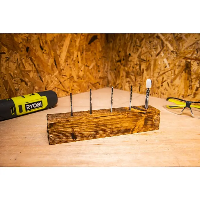 Ryobi Bohrersatz RAR404-4 - 4 Stk. - Verschiedene Bohrer sind in einem Holzblock als Halterung platziert, zusammen mit einer gelben Bohrmaschine und einer Schutzbrille.