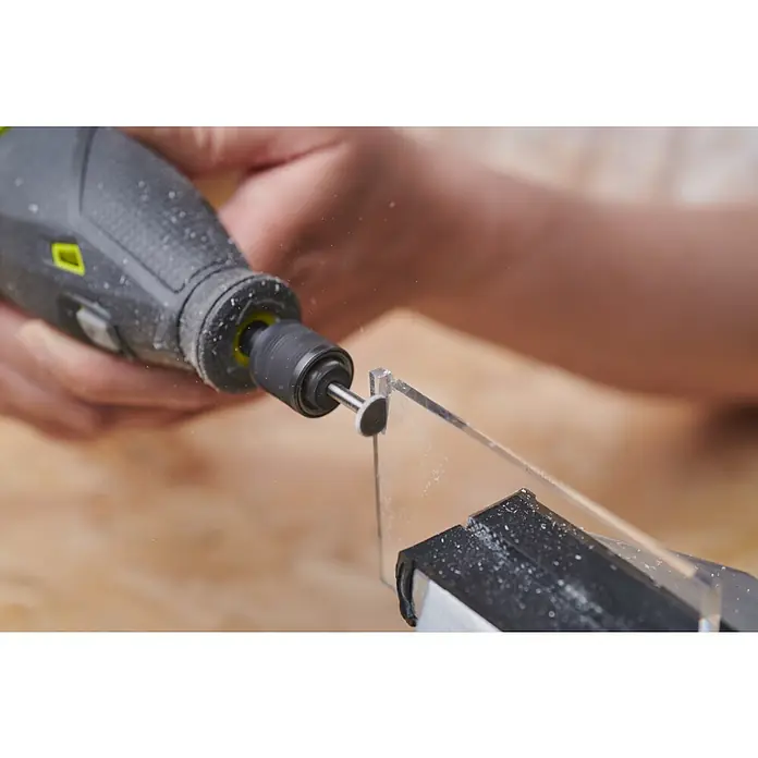 Ryobi Fräsmesser RAR103 rund - Arbeitsdurchmesser: 9,5 mm, Passend für: Rotationswerkzeuge mit 3,2 mm (1/8") Spannzangenkapazität - Die Nahaufnahme zeigt ein Präzisionswerkzeug beim Bearbeiten eines transparenten Kunststoffstücks, das in einem Schraubstock fixiert ist. Späne fliegen, was auf eine detaillierte Arbeit hindeutet.