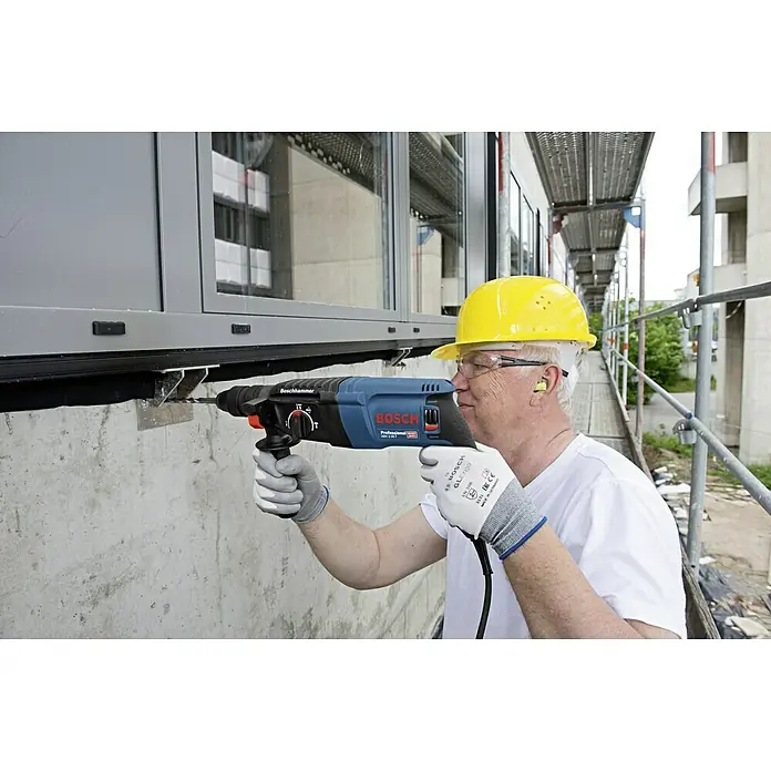 Bosch Professional Martillo combinado GBH 2-26 F - 830 W, 2,7 J, Alojamiento SDS-Plus - Un trabajador de la construcción usa un martillo perforador Bosch para hacer un agujero en una pared de hormigón. Casco y gafas protegen.