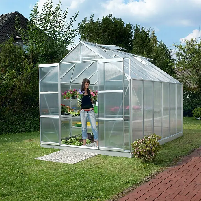 Vitavia Gewächshaus Mars 9900 - Außenmaß inkl. Dachüberstand (B x T): 257 x 383 cm, 4 mm, Aluminium - Eine Frau betritt ein Gewächshaus aus Aluminium, das im Garten steht und Pflanzen beherbergt. Das Gewächshaus ist lichtdurchlässig und stabil.