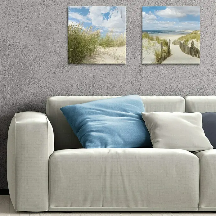 ProArt Maritime Glasbild - Sommertag II, B x H: 50 x 50 cm - Nahaufnahme eines modernen Sofas mit blauen und weißen Kissen vor einer Wand mit zwei gerahmten Strandbildern.