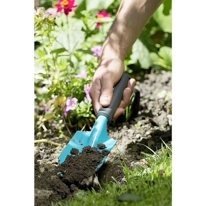 Gardena Classic Blumenkelle - Stahl - Nahaufnahme einer Person, die mit einer blauen Gartenschaufel Erde aushebt, um ein Blumenbeet vorzubereiten.