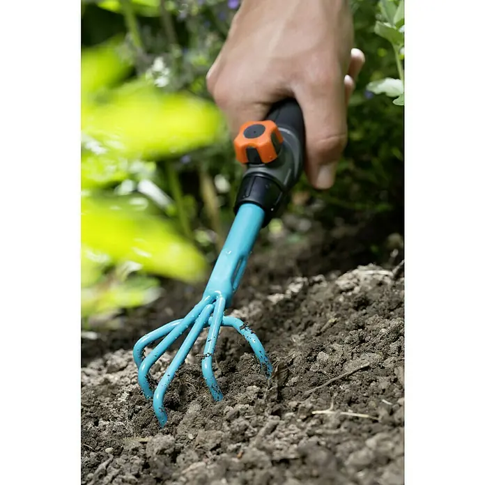 Gardena Combisystem Blumenkralle - Arbeitsbreite: 7 cm, Stahl - Nahaufnahme einer Hand, die eine blaue Gartenkralle mit orangem Knopf in dunkle Erde drückt, um den Boden aufzulockern.