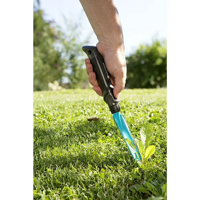 Gardena Unkrautstecher - 14,5 cm - Ein Unkrautstecher von Gardena wird in einer grünen Rasenfläche verwendet, um Unkraut zu entfernen. Das Werkzeug hat einen blauen Schaft und schwarzen Griff.