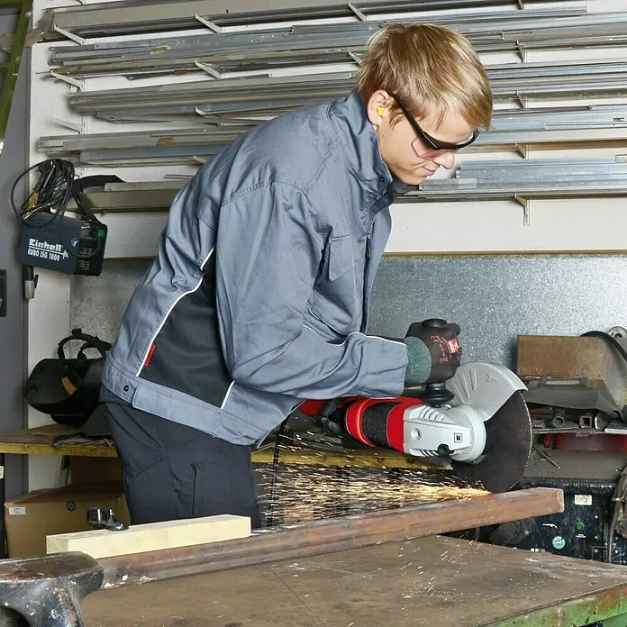 Einhell Expert Elektro-Winkelschleifer TE-AG 230/2000 - 2.000 W, 230 mm, Leerlaufdrehzahl: 6.500 U/min - Ein Mann in Arbeitskleidung schleift ein Metallstück mit einem Winkelschleifer, wobei Funken sprühen. Er trägt Schutzbrille und Gehörschutz für die Arbeitssicherheit.