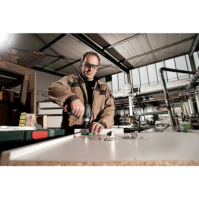 Metabo Akku-Bohrschrauber-Set PowerMaxx BS Basic - 12 V, 2 Akkus, 2 Ah, 34 Nm, 41 -tlg. - Ein Handwerker mit Schutzbrille befestigt Beschläge mit einem Metabo Akku-Bohrschrauber an einem Werkstück. Perfekt für präzise Montagearbeiten.
