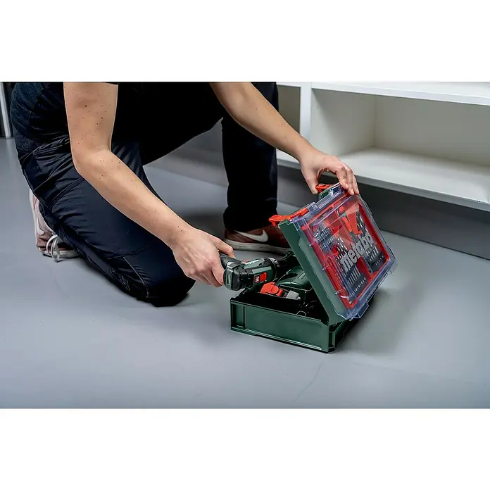 Metabo Akku-Bohrschrauber-Set PowerMaxx BS Basic - 12 V, 2 Akkus, 2 Ah, 34 Nm, 41 -tlg. - Ein Mann kniet und öffnet einen Metabo Werkzeugkoffer mit einem Akkuschrauber und einer Bohrer-Bit-Sammlung.