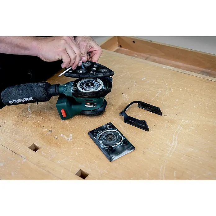 Metabo Akku-Multischleifer PowerMaxx SMA 12 BL - 12 V, Ohne Akku, Breite Schleifplatte: 100 mm, Länge Schleifplatte: 150 mm - Auseinandergebauter Metabo Kombischleifer mit verschiedenen Schleifplatten und Staubabsaugung auf einer Werkbank.