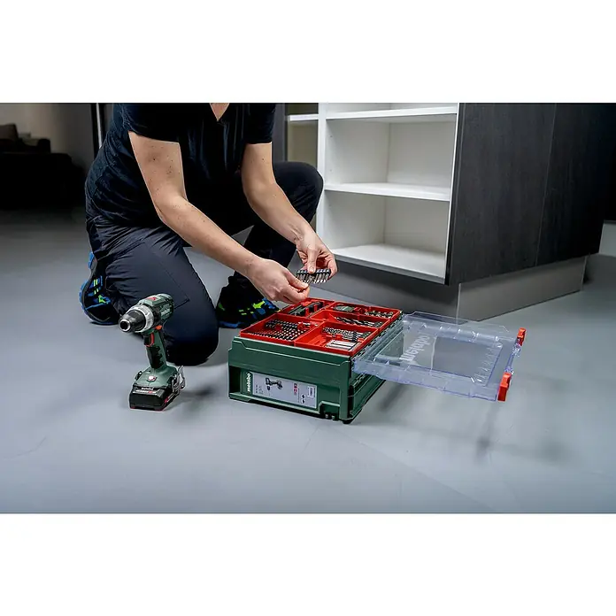 Metabo CAS 18V Akku-Schlagbohrschrauber-Set SB 18 - 18 V, 2 Akkus, 2 Ah, 87 -tlg. - Eine Person, die vor einem Metabo-Werkzeugkasten mit Bohraufsätzen kniet, mit einem Akkuschrauber daneben.