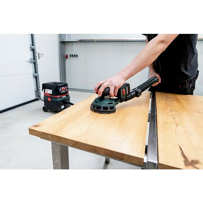 Metabo Akku-Exzenterschleifer PowerMaxx SXA 12-125 BL - 12 V, Ohne Akku - Metabo Exzenterschleifer im Einsatz: Ein Handwerker schleift eine Holzplatte mit dem leistungsstarken Schleifgerät, angeschlossen an einen Staubsauger.