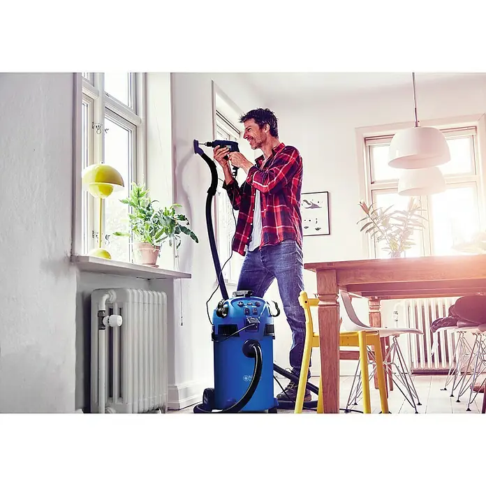 Nilfisk Elektro-Nass- & Trockensauger Multi II 30 T EU - 1.400 W, Behältervolumen: 30 l, Halbautomatisch - Ein Mann bohrt mit einem Nilfisk Nass- und Trockensauger an der Wand, um den Staub aufzufangen. Das Gerät ist blau und hat einen schwarzen Schlauch.
