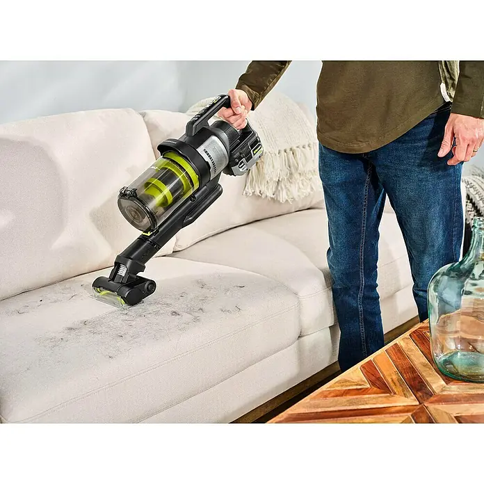 Ryobi ONE+ Akku-Staubsauger RASV18BL-0 - 18 V, Ohne Akku, Behältervolumen: 1.000 ml - Bild einer Person, die mit einem kabellosen Staubsauger ein cremefarbenes Sofa von Tierhaaren befreit. Der Staubsauger ist schwarz mit grünen Akzenten.