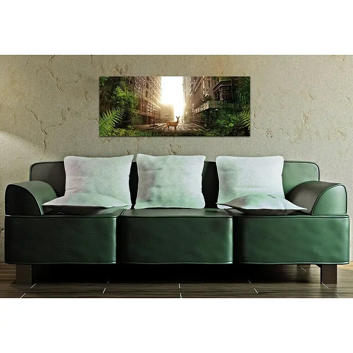 Glasbild - Das Reh, B x H: 125 x 50 cm - Ein modernes Wohnzimmer mit einer grünen Ledercouch und einem Wandbild, das eine von Natur überwucherte Stadt mit einem Reh zeigt.