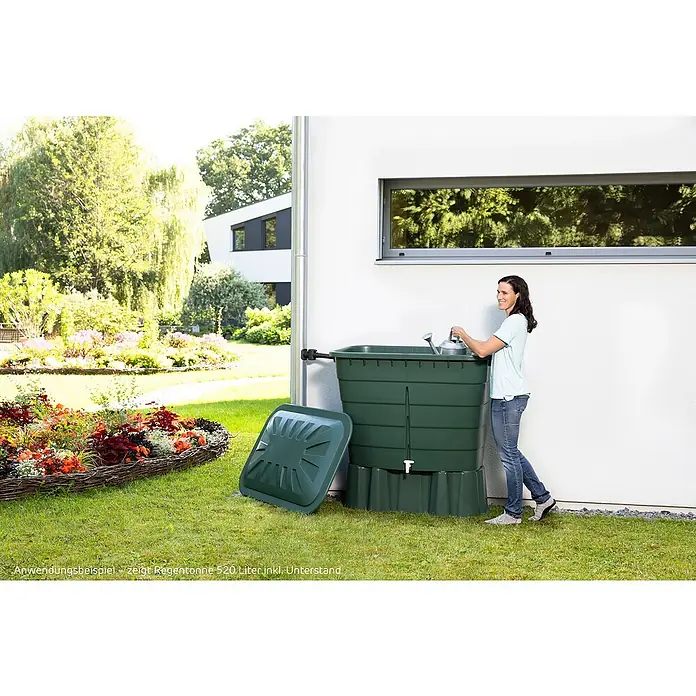 Garantia Basic Line Regentonne - 520 l, Grün, 1 Stk. - Frau befüllt Regentonne im Garten. Die Regentonne ist grün und steht an einer weißen Hauswand neben einem Blumenbeet.