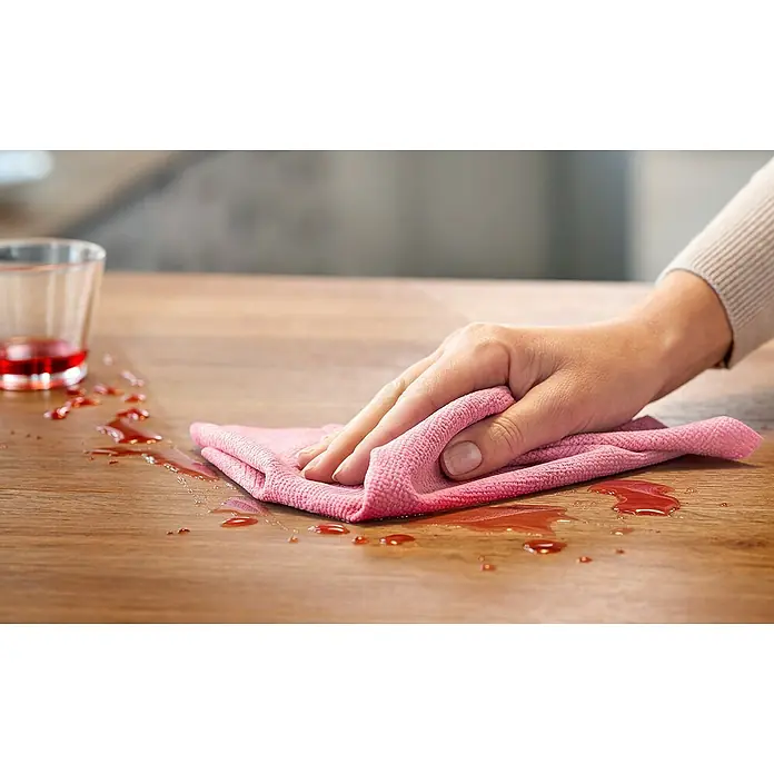 Vileda Universalreinigungstücher Actifibre Soft Touch - 2 Stk., Mikrofaser - Ein Tisch wird mit einem rosa Tuch abgewischt, um verschüttete Flüssigkeit zu entfernen, mit einem Glas im Hintergrund.