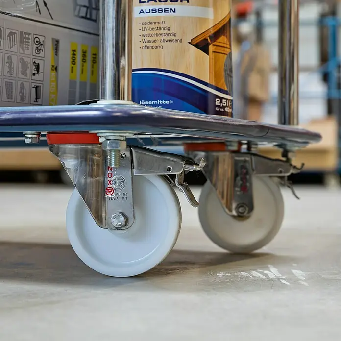 Dörner & Helmer Transportgeräterolle - Durchmesser Rollen: 100 mm, Traglast: 150 kg, Gleitlager, Mit Platte und Feststeller - Nahaufnahme von Rollen eines Transportwagens mit Feststellbremsen, der mit Produkten wie Lack beladen ist.