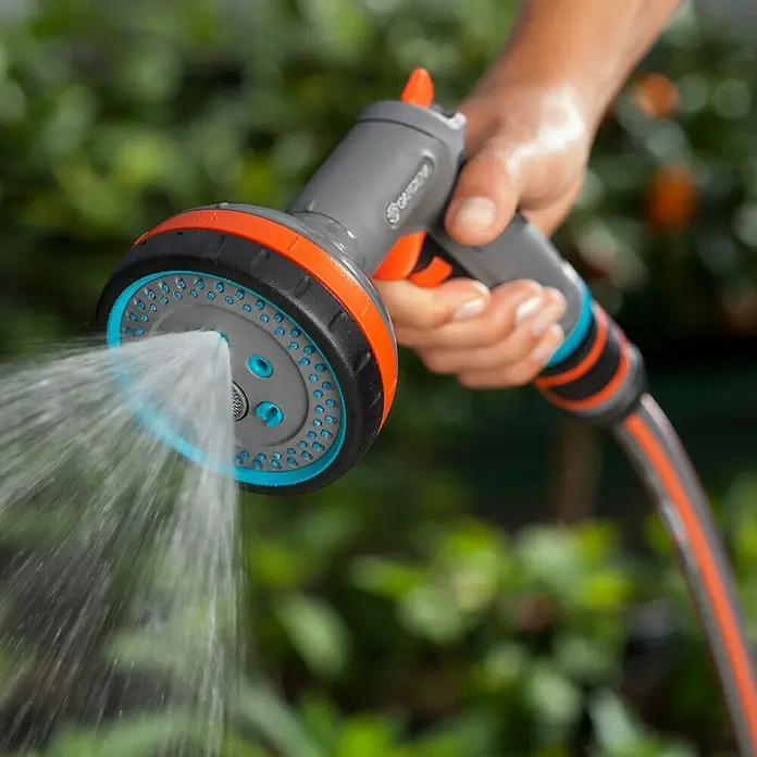 Gardena Comfort Multibrause - Anzahl Sprühformen: 5 Stk., Metall/Kunststoff - Eine Gardena Mehrzweckbrause wird von einer Hand gehalten und sprüht Wasser. Sie ist grau, orange, schwarz und blau und an einen Gartenschlauch angeschlossen.