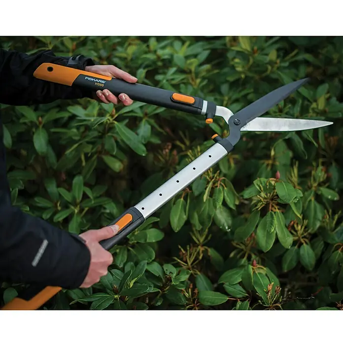Fiskars Smartfit Hand-Heckenschere HS86 - 68 cm - 93 cm - Eine Person benutzt eine Fiskars HS56 Gartenschere vor einer grünen Hecke. Die Schere hat orangefarbene Akzente und ausziehbare Griffe.
