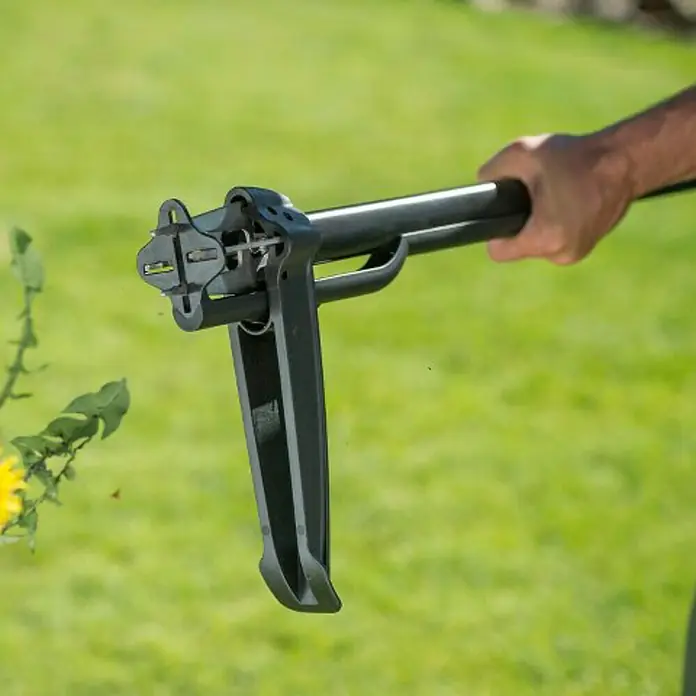 Fiskars Xact Unkrautstecher - Mit Greifarmen, Länge: 100 cm - Nahaufnahme einer schwarzen Unkrautwurzelentfernungswerkzeug im Einsatz vor einem grünen Rasen.