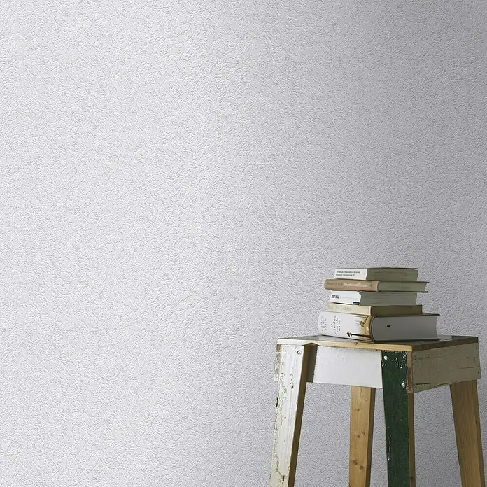 Rasch Papiertapete Struktur - Weiß, Uni, 20 x 0,53 m - Detailaufnahme einer weißen, strukturierten Tapete mit einem Stapel Büchern auf einem kleinen Hocker im Vordergrund.