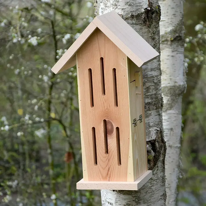 Dobar Schmetterlingsunterschlupf Sonnenfalter - 23 x 14 x 38 cm, Kiefer - Ein hölzernes Insektenhotel mit Schlitzen zur Nisthilfe hängt an einem Baum. Das naturbelassene Holz fügt sich in die natürliche Umgebung ein.