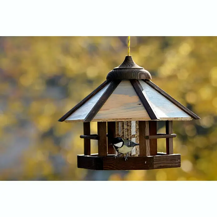Dobar Sechseck-Vogelfutterhaus Camouflage - 35 x 35 x 29 cm, Ausstattung: Mit Silo - Ein hölzernes Vogelhaus hängt an einem gelben Seil, während ein Vogel darin sitzt und sich an den Körnern labt.