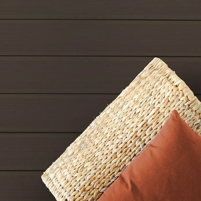 Kovalex WPC-Terrassendiele Massiv strukturo nach Maß - 300 x 14,5 x 2 cm, Schoko - Nahaufnahme eines Korbsessels mit einem orangefarbenen Kissen auf einem Hintergrund aus horizontalen braunen Holzpaneelen.