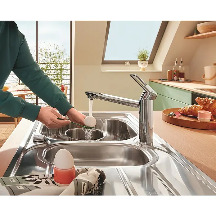 Grohe QuickFix Start Loop Spültischarmatur - Ausladung Armatur: 222 mm, Chrom, Hochglänzend - Ein Edelstahl-Spültisch mit einer modernen Armatur, an der eine Person ein Ei unter fließendem Wasser reinigt, umgeben von Frühstückszutaten.
