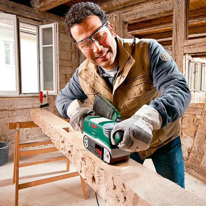 Bosch Lijadora de banda eléctrica de sobremesa PBS 75 AE - 750 W - Un hombre lija una tabla de madera con una lijadora de banda verde Bosch. Lleva gafas de seguridad, guantes y un chaleco.