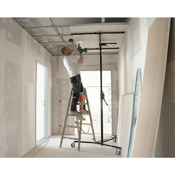 Rikta Plattenlift - 40 kg, Höhenverstellung: 155 cm - 300 cm - Ein Mann montiert eine Gipskartondecke mit einem Akkuschrauber, stehend auf einer Leiter. Werkzeuge und Baumaterialien sind im Raum verteilt.