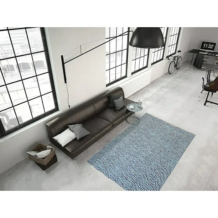 Kayoom Flachgewebeteppich Cocktail 400 - Blau, 230 x 160 cm, 100% Baumwolle - Wohnzimmeransicht mit blau gemustertem Teppich, braunem Sofa und großen Fenstern, die viel Licht hereinlassen. Stilvolles und modernes Design.