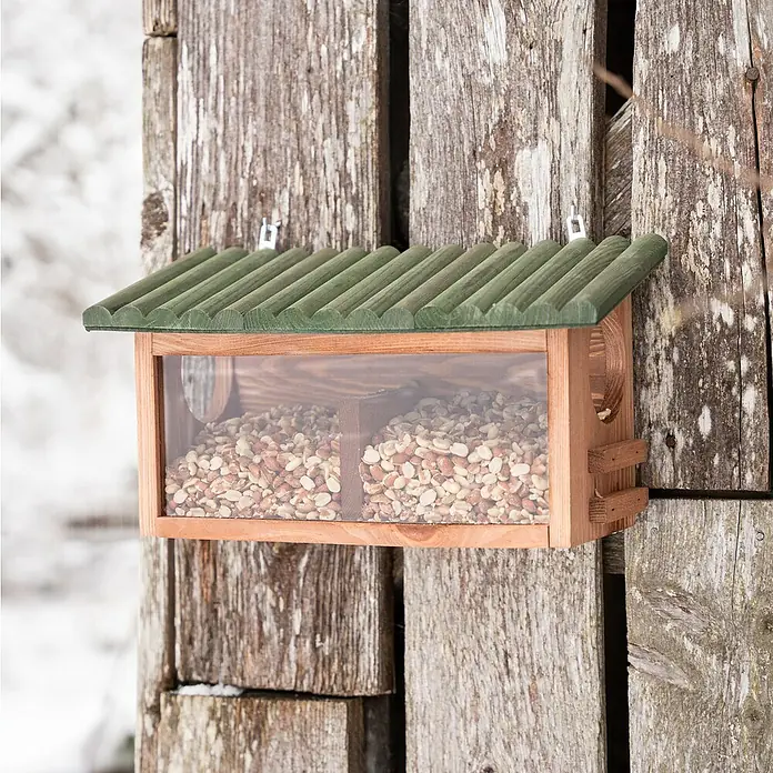 Windhager Futterstation XL - Holz, Braun/Grün - Ein hölzernes Vogelhaus mit einem grünen Dach, gefüllt mit Erdnüssen, hängt an einer hölzernen Wand. Ideal für Gartenliebhaber und Vogelfreunde.