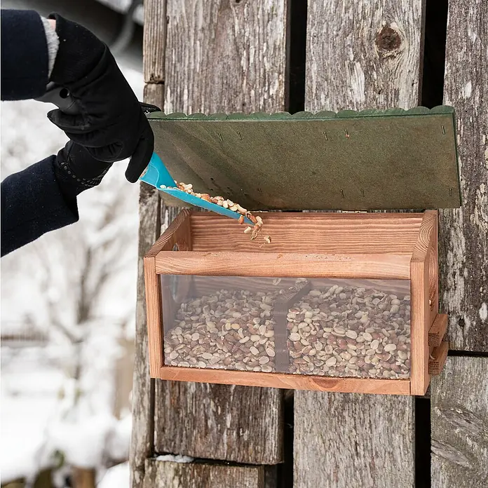 Windhager Futterstation XL - Holz, Braun/Grün - Eine Person befüllt einen hölzernen Vogelhäuschen mit Erdnüssen. Das Vogelhaus ist an einer Holzwand befestigt und mit Nüssen gefüllt.