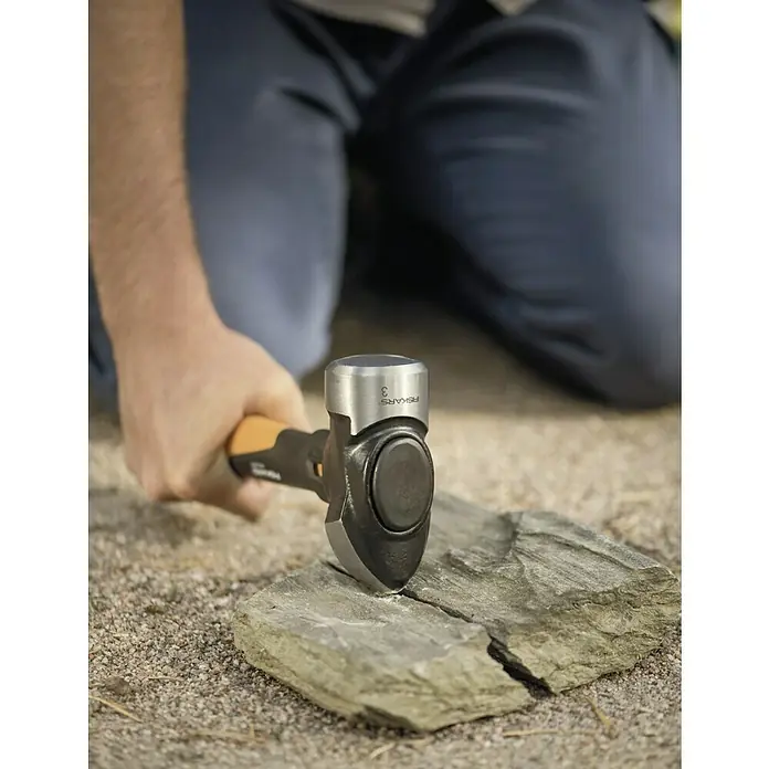 Fiskars Fäustel L - Gewicht Kopf: 1.800 g, Material Stiel: Glasfaserverstärkter Kunststoff (GfK) - Nahaufnahme eines Mannes, der mit einem Fiskars Spalthammer einen Stein spaltet, was die Werkzeugqualität hervorhebt.