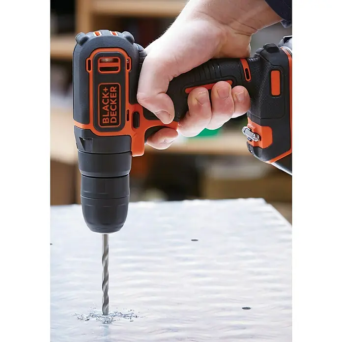 Black+Decker Taladro atornillador percutor de batería BDCHD18KB - 18 V, 2 baterías, 1,5 Ah, 40 Nm - Un taladro inalámbrico Black+Decker se utiliza para perforar un material de color claro, con virutas de perforación acumulándose en la superficie.